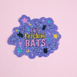 Puede incluir: Una pegatina hologr&aacute;fica morada con forma de nube y el texto "It's Frickin' Bats" en letras rosas y amarillas. La pegatina est&aacute; decorada con murci&eacute;lagos negros, estrellas y lunas.
