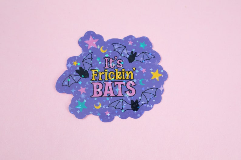 Puede incluir: Una pegatina hologr&aacute;fica morada con forma de nube y el texto "It's Frickin' Bats" en letras rosas y amarillas. La pegatina est&aacute; decorada con murci&eacute;lagos negros, estrellas y lunas.