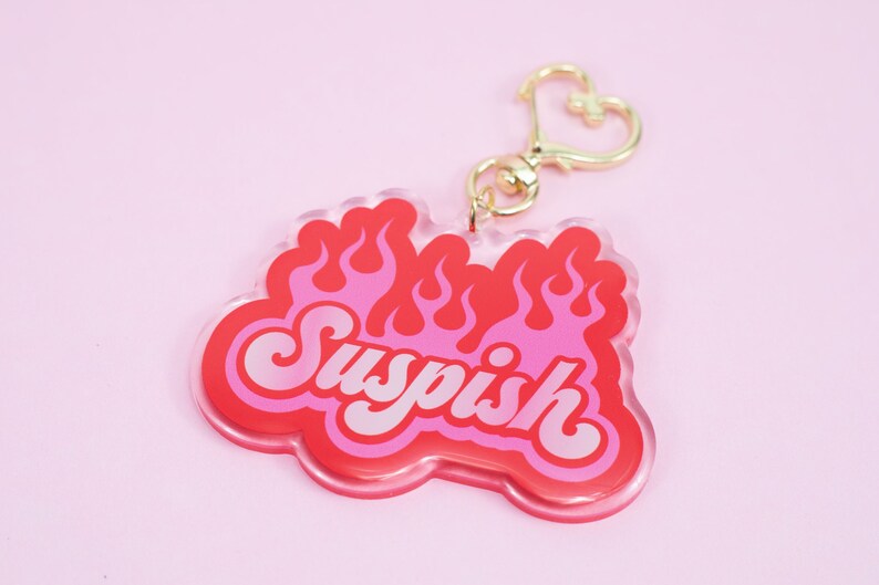 Pode incluir: Um chaveiro de acr&iacute;lico rosa e vermelho com a palavra "Suspish" em uma fonte retr&ocirc;, cercado por chamas. O chaveiro tem um fecho dourado em forma de cora&ccedil;&atilde;o.