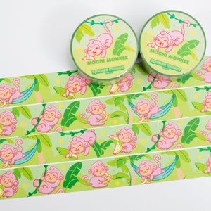 Könnte beinhalten: Zwei Rollen grünes und weißes Washi-Tape mit einem rosafarbenen Affen-Design. Das Band zeigt einen Affen, der eine Banane hält, einen Affen, der an einer Rebe schwingt, und einen Affen, der in einer Hängematte schläft. Das Band ist mit "Mochi Monkee" und "Sprinkle Monkee" beschriftet.
