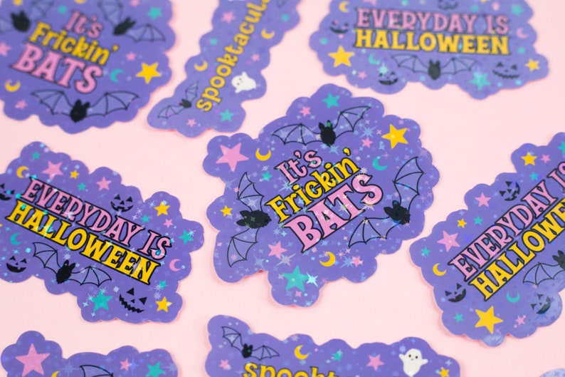Puede incluir: Pegatinas de Halloween moradas y amarillas con purpurina y varios dise&ntilde;os. Las pegatinas presentan murci&eacute;lagos, estrellas, lunas y el texto "It's Frickin' Bats" y "Everyday is Halloween".