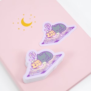 May include: Two stickers featuring a black cat sleeping on a book with a pumpkin and a lollipop. The stickers are on a pink background with a gold moon and stars.
