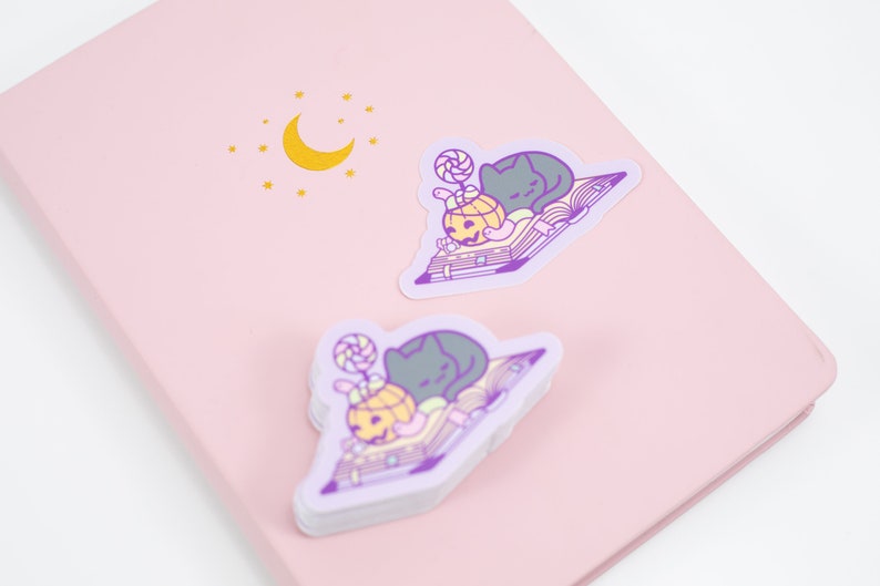 May include: Two stickers featuring a black cat sleeping on a book with a pumpkin and a lollipop. The stickers are on a pink background with a gold moon and stars.