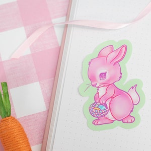 May include: A pink bunny rabbit sticker with a basket of colorful Easter eggs. The bunny is holding the basket in its front paws and has a sad expression on its face. The sticker is on a green background.