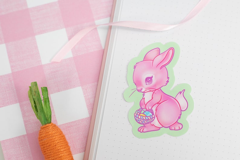 May include: A pink bunny rabbit sticker with a basket of colorful Easter eggs. The bunny is holding the basket in its front paws and has a sad expression on its face. The sticker is on a green background.