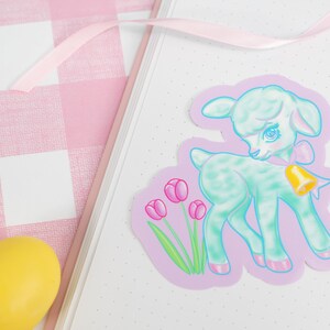 May include: A pastel green lamb sticker with a pink bow and a gold bell. The lamb is standing on a purple background with pink tulips.