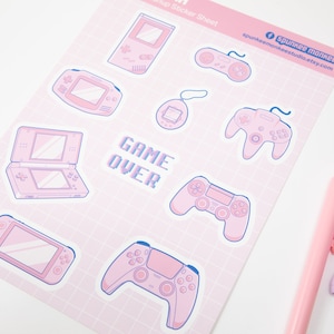 May include: A sheet of pink and blue video game-themed stickers. The stickers feature various game consoles and controllers, along with the text "GAME OVER." The sheet has a grid pattern and the brand name "spunkee monkee" is visible.