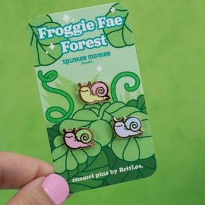 May include: Three gold enamel pins featuring cartoon snails with different coloured shells. The snails are on a green card with the text "Froggy Fae Forest" and "Spunkee Monkee Studio" and "enamel pins by BritLos."