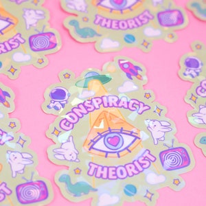 May include: A set of holographic stickers with a green background and a variety of pastel colors. The stickers feature a pyramid, an eye, a UFO, a rocket, a dinosaur, a bunny, a television, and the text "Conspiracy Theorist".
