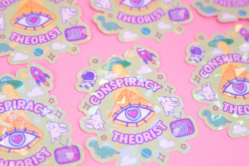 May include: A set of holographic stickers with a green background and a variety of pastel colors. The stickers feature a pyramid, an eye, a UFO, a rocket, a dinosaur, a bunny, a television, and the text "Conspiracy Theorist".