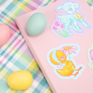 May include: A pink notebook with four pastel colored stickers of baby animals. The stickers feature a lamb, a chick, a bunny, and a flower. The notebook is on a pink and green plaid surface with three pastel colored Easter eggs.