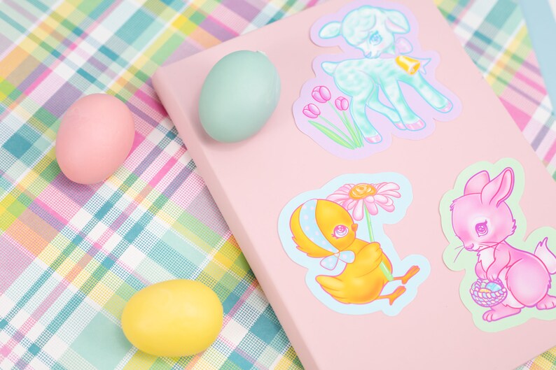 May include: A pink notebook with four pastel colored stickers of baby animals. The stickers feature a lamb, a chick, a bunny, and a flower. The notebook is on a pink and green plaid surface with three pastel colored Easter eggs.