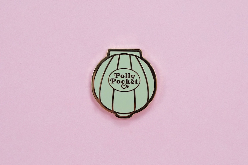 Puede incluir: Un pin de esmalte Polly Pocket verde menta con borde dorado. El pin presenta las palabras "Polly Pocket" en un dise&ntilde;o circular con un s&iacute;mbolo de coraz&oacute;n. El pin est&aacute; sobre un fondo rosa.