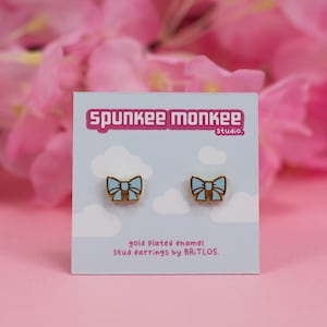 Könnte beinhalten: Ein Paar goldplattierte Emaille-Ohrstecker mit einem hellblauen Schleifendesign. Die Ohrringe werden auf einer Karte mit dem Text "Spunkee Monkee Studio" und "goldplattierte Emaille-Ohrstecker von BRITLOS." angezeigt.