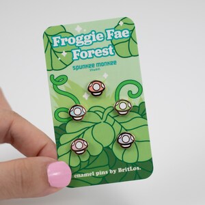 K&ouml;nnte beinhalten: Satz mit vier Emaille-Pins mit verschiedenen farbigen Pilzen mit wei&szlig;en Punkten. Die Pins befinden sich auf einer Karte mit gr&uuml;nem Hintergrund und dem Text "Froggy Fae Forest" und "enamel pins by BritLos."