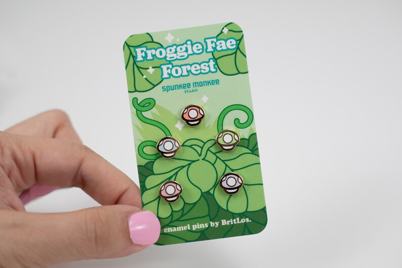 K&ouml;nnte beinhalten: Satz mit vier Emaille-Pins mit verschiedenen farbigen Pilzen mit wei&szlig;en Punkten. Die Pins befinden sich auf einer Karte mit gr&uuml;nem Hintergrund und dem Text "Froggy Fae Forest" und "enamel pins by BritLos."