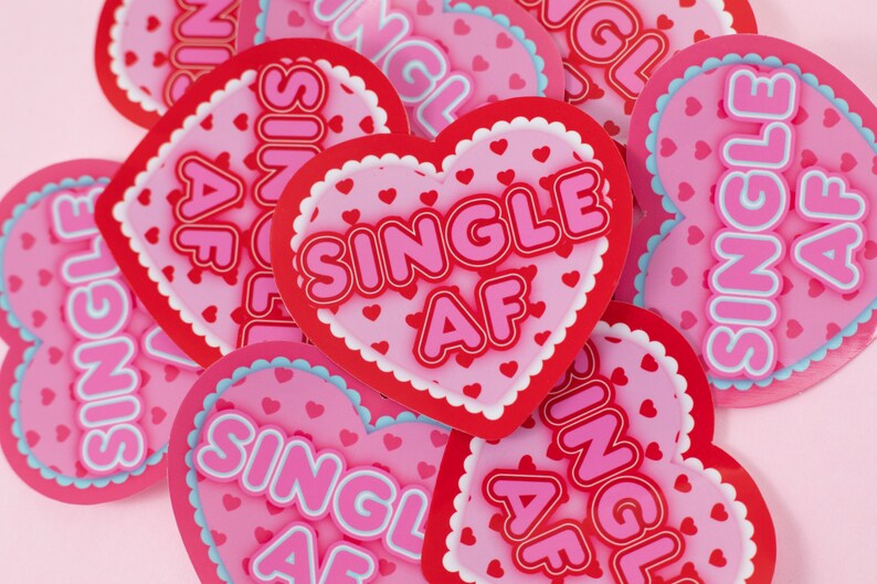 Op de afbeelding: Een stapel roze en rode hartvormige stickers met de tekst "Single AF" erop. De stickers hebben een witte omtrek en zijn versierd met kleine roze hartjes.