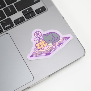 May include: A purple and white sticker with a black cat sleeping on a book. The book has a yellow pumpkin with a jack-o-lantern face and a pink and white lollipop on top.
