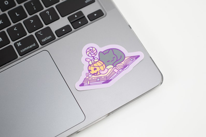 May include: A purple and white sticker with a black cat sleeping on a book. The book has a yellow pumpkin with a jack-o-lantern face and a pink and white lollipop on top.