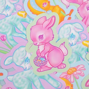 May include: A collection of pastel-colored Easter stickers featuring a pink bunny holding a basket of eggs, a blue lamb, and a yellow chick. The stickers are on a light blue background.