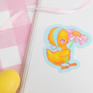 May include: A yellow chick with a blue polka dot bow holding a pink flower. The chick is on a blue background with white polka dots.