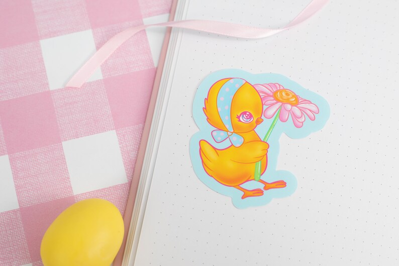 May include: A yellow chick with a blue polka dot bow holding a pink flower. The chick is on a blue background with white polka dots.
