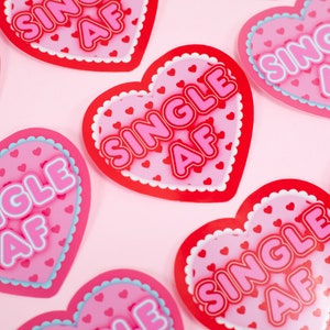Op de afbeelding: Een set van negen roze en rode hartvormige stickers met de tekst "SINGLE AF" in witte en roze letters. De stickers zijn versierd met kleine roze hartjes.