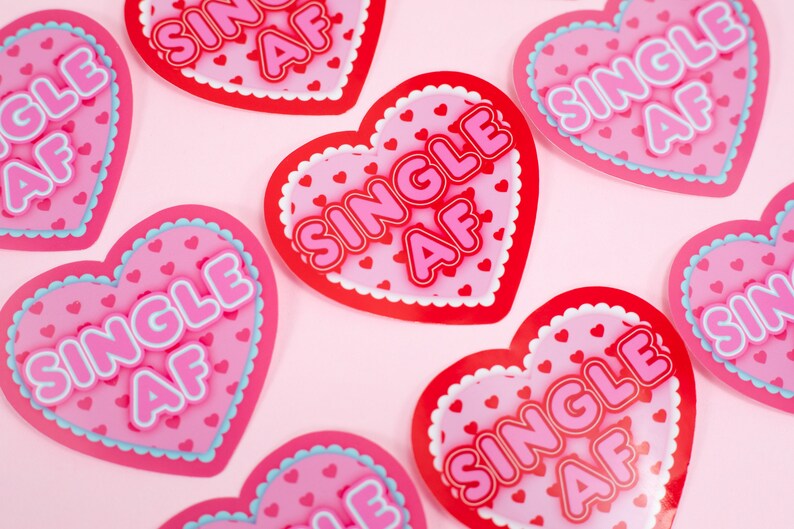 Op de afbeelding: Een set van negen roze en rode hartvormige stickers met de tekst "SINGLE AF" in witte en roze letters. De stickers zijn versierd met kleine roze hartjes.