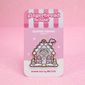May include: Pink gingerbread house enamel pin with candy cane details, frosting, and a heart-shaped door. The pin is on a pink card with the text "gingerbread house", "spunkee monkee studio", and "enamel pins by BRiTLOS."