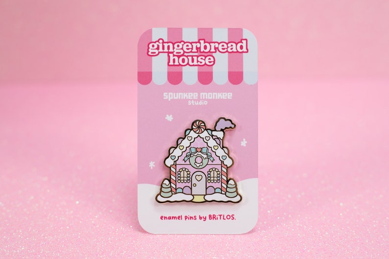 May include: Pink gingerbread house enamel pin with candy cane details, frosting, and a heart-shaped door. The pin is on a pink card with the text "gingerbread house", "spunkee monkee studio", and "enamel pins by BRiTLOS."