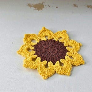 Op de afbeelding: Een handgemaakte gehaakte zonnebloem met een bruin hart en gele bloemblaadjes. De zonnebloem is een decoratief item, waarschijnlijk een onderzetter of applicatie, op een witte ondergrond.