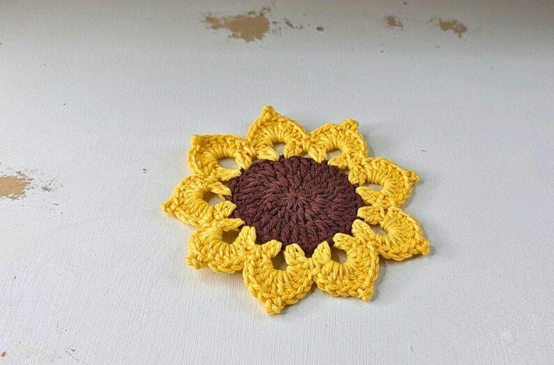 Op de afbeelding: Een handgemaakte gehaakte zonnebloem met een bruin hart en gele bloemblaadjes. De zonnebloem is een decoratief item, waarschijnlijk een onderzetter of applicatie, op een witte ondergrond.