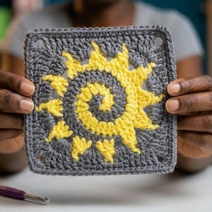 May include: A crocheted square with a yellow sun motif on a grey background. The sun design has a spiral centre and radiating rays. The piece is held up, highlighting the handmade details and colour contrast.