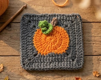 May include: A square crochet granny square featuring a dark gray background with a centered orange pumpkin motif. The pumpkin has a small green crocheted leaf and a brown stem attached to the top center of the orange circular shape.