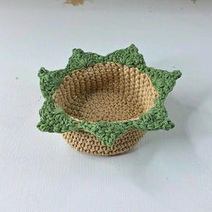 Op de afbeelding: Een handgemaakte gehaakte mand met een stervormige groene rand en een bruin interieur. De mand is rond en heeft een getextureerd uiterlijk, perfect voor het vasthouden van kleine items. De mand heeft een diameter van ongeveer 15 cm.