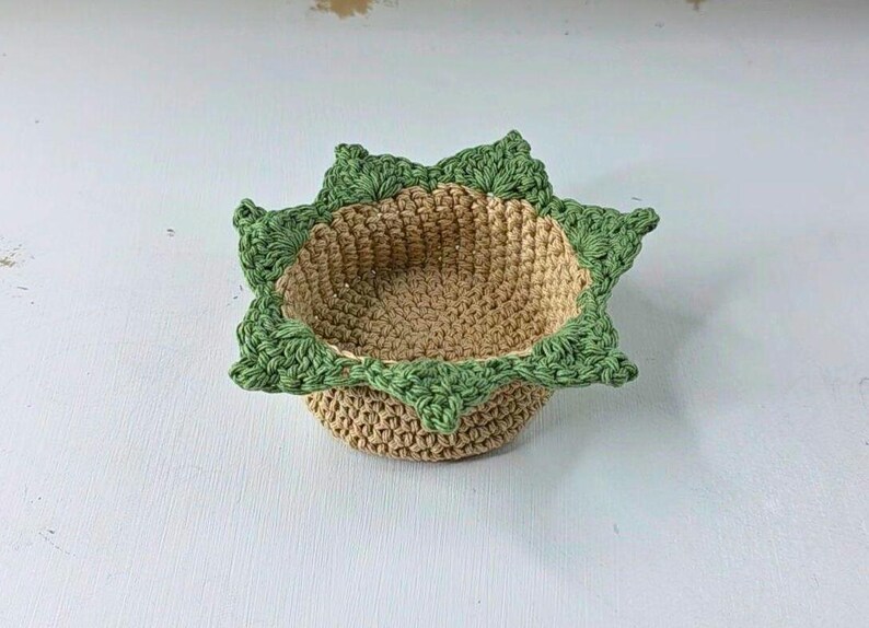 Op de afbeelding: Een handgemaakte gehaakte mand met een stervormige groene rand en een bruin interieur. De mand is rond en heeft een getextureerd uiterlijk, perfect voor het vasthouden van kleine items. De mand heeft een diameter van ongeveer 15 cm.