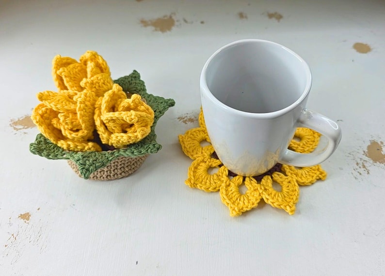 Op de afbeelding: Een witte keramische mok staat op een gele gehaakte zonnebloemonderzetter. Daarnaast staat een klein gehaakt mandje met gele gehaakte bloemen met groene bladeren.