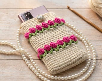 May include: A beige crochet phone pouch featuring two horizontal rows of pink tulip appliqués with green stems. The pouch includes a white pearl bead strap and a single clear rhinestone button closure at the top, holding a smartphone.
