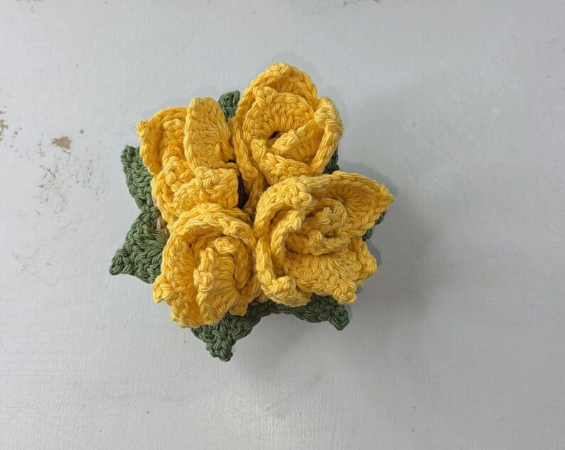 Op de afbeelding: Een close-up van een handgemaakt gehaakt bloemstuk. Het toont heldergele rozen en groene bladeren, allemaal ingewikkeld vervaardigd. Het gestructureerde, driedimensionale ontwerp van de bloemen voegt een vleugje handgemaakte charme toe. Ideaal om te decoreren.