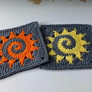 以下が含まれることがあります： 太陽のデザインが施された、2つの正方形のかぎ針編み作品。1つは灰色の背景にオレンジ色の太陽、もう1つは黄色の太陽が特徴です。太陽は渦巻きの中心と尖った光線を持っています。かぎ針編みの作品は詳細です。