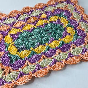 May include: A handmade, oval-shaped crochet doily in a vibrant array of colors. The design features a central teal spiral, surrounded by yellow, purple, and orange fan-shaped patterns. The edges are finished with a scalloped border.