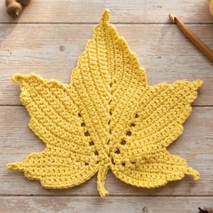 May include: A bright yellow crocheted maple leaf with a textured surface and openwork details along the central veins. The leaf is crafted from yarn and features a pointed, multi-lobed shape with a small stem at the base.