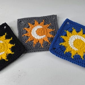 May include: Three crocheted squares with celestial designs. One is black with a yellow sun and white crescent moon, another is gray with an orange sun and white moon, and the third is blue with a yellow sun and white moon.