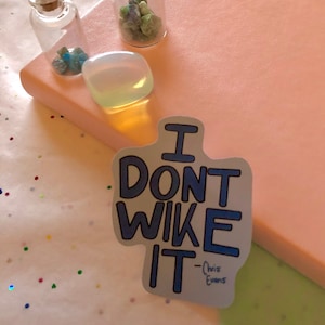 以下が含まれることがあります： 薄い灰色の背景に「I don't like it - Chris Evans」というテキストが書かれたステッカー。ステッカーは、緑と青のクリスタルで満たされた2つの小さなガラス瓶のあるピンクのノートブックの上に置かれています。