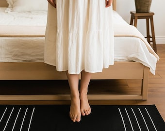 May include: A black rectangular rug with white diagonal lines. The rug is placed on a wooden floor in front of a bed. A person wearing a white dress is standing on the rug.