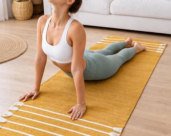 May include: A rectangular mustard yellow yoga mat featuring a textured surface with white horizontal stripes and decorative white tassels at both ends. The mat is laid flat on a light-colored wooden floor, providing a stable surface for exercise.