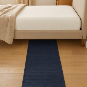 K&ouml;nnte beinhalten: Ein rechteckiger, marineblauer Teppich liegt auf einem hellen Holzboden unter einem Bett. Der Teppich hat eine strukturierte Oberfl&auml;che und befindet sich vor dem Bett. Das Bett hat ein beiges Kopfteil und eine wei&szlig;e Matratze.