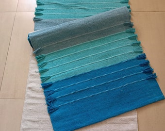 May include: A rectangular woven runner rug featuring a gradient of blue and teal horizontal stripes. The rug has a textured weave with small decorative tassels along the side edges. The bottom section transitions into a solid off-white color.