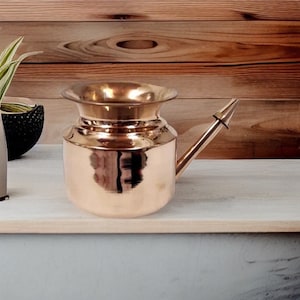 May include: A copper-colored neti pot with a wide rim and a spout. The pot is made of shiny metal and reflects the surrounding environment. The neti pot is designed for nasal irrigation.