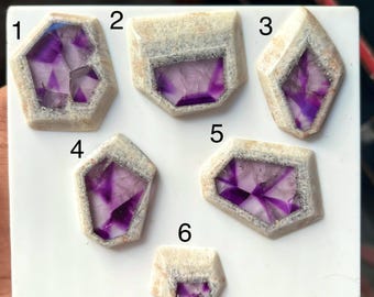 Puede incluir: Seis piedras de formas geométricas irregulares con un borde exterior beige texturizado y un centro cristalino de color púrpura intenso translúcido. Cada piedra está etiquetada con un número negro del uno al seis sobre una base blanca.
